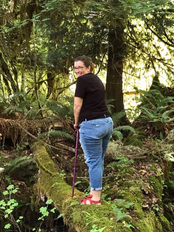 A head and shoulders photo of Angie Ebba, a woman with ankylosing spondylitis, fibromyalgia and migraine. She is standing in a forrested area, leaning on her walking stick and turning her head back to smile at the camera.